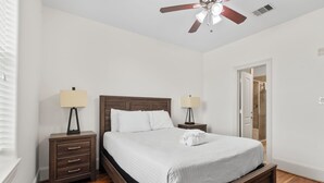 Un dormitorio con una cama de madera, dos mesitas de noche con lámparas, un ventilador de techo y una puerta que da a otra habitación.