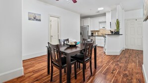 Un comedor con una mesa y sillas de madera, un ventilador de techo y una cocina al fondo.