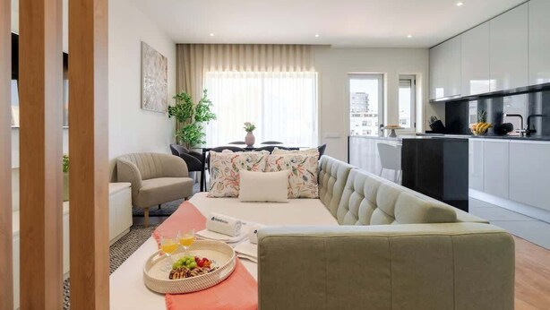 Een moderne woonkamer met een bank, een eettafel met fruit en een dienblad, een keuken met kasten en een uitzicht op de stad door grote ramen.