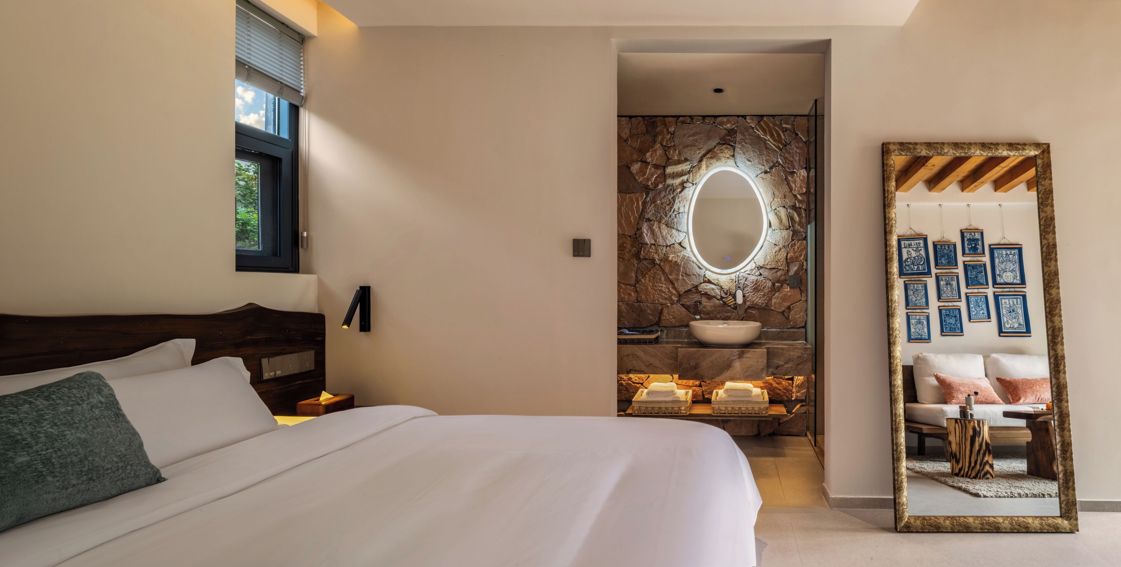 A bedroom with a bed, a nightstand, a mirror, a stone wall with a sink, and a bathroom with a towel rack.