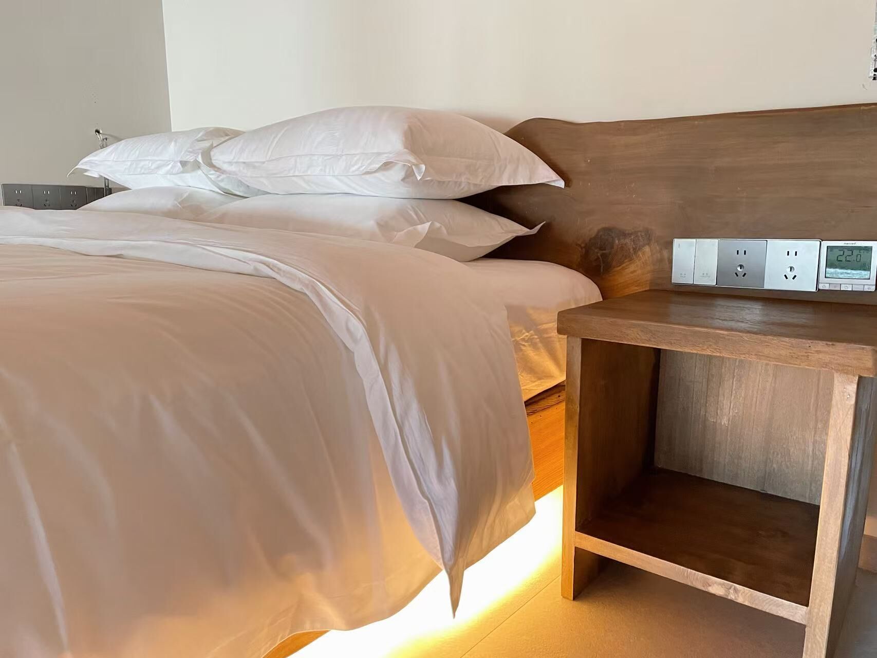 A neatly made bed with white linens and a wooden headboard. There is a bedside table with a power outlet and a digital clock displaying the time.