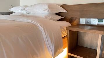 A neatly made bed with white linens and a wooden headboard. There is a bedside table with a power outlet and a digital clock displaying the time.
