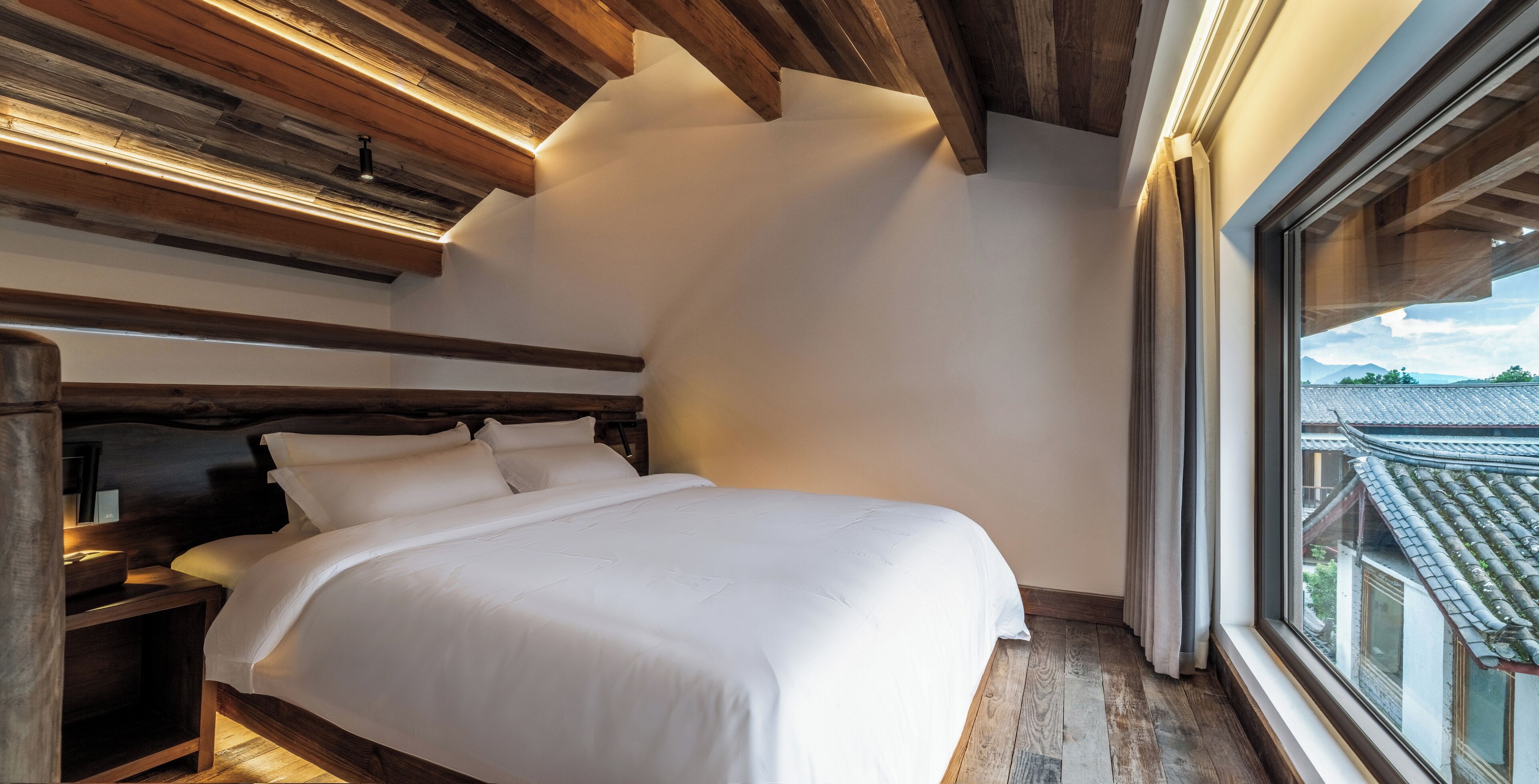 A bedroom with a wooden headboard, a large bed with white bedding, and a view of traditional architecture through a window.
