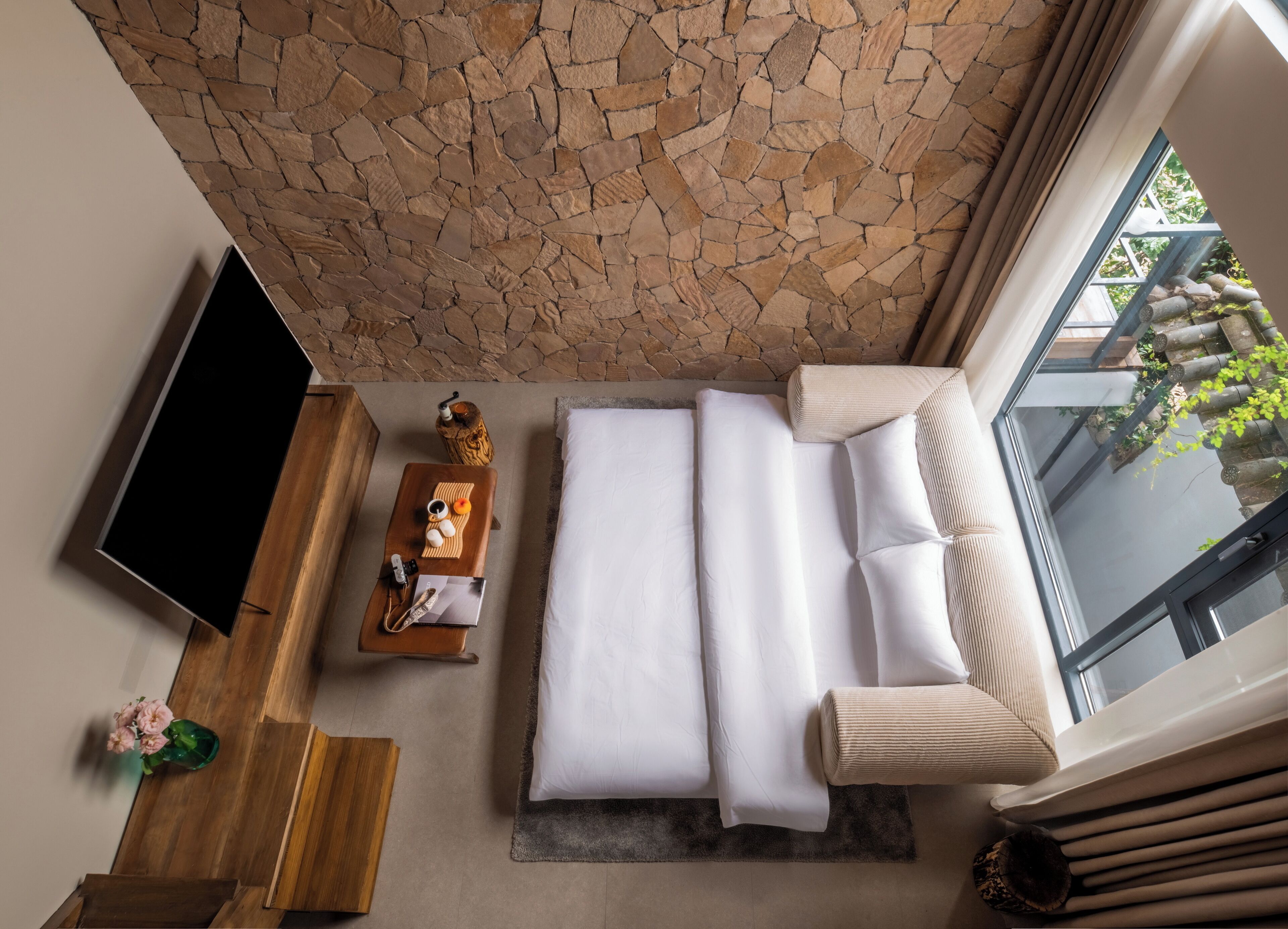 A modern hotel room with a stone wall, a flat-screen TV, a wooden side table with toiletries, a sofa with white bedding, and a large window offering a view of greenery.