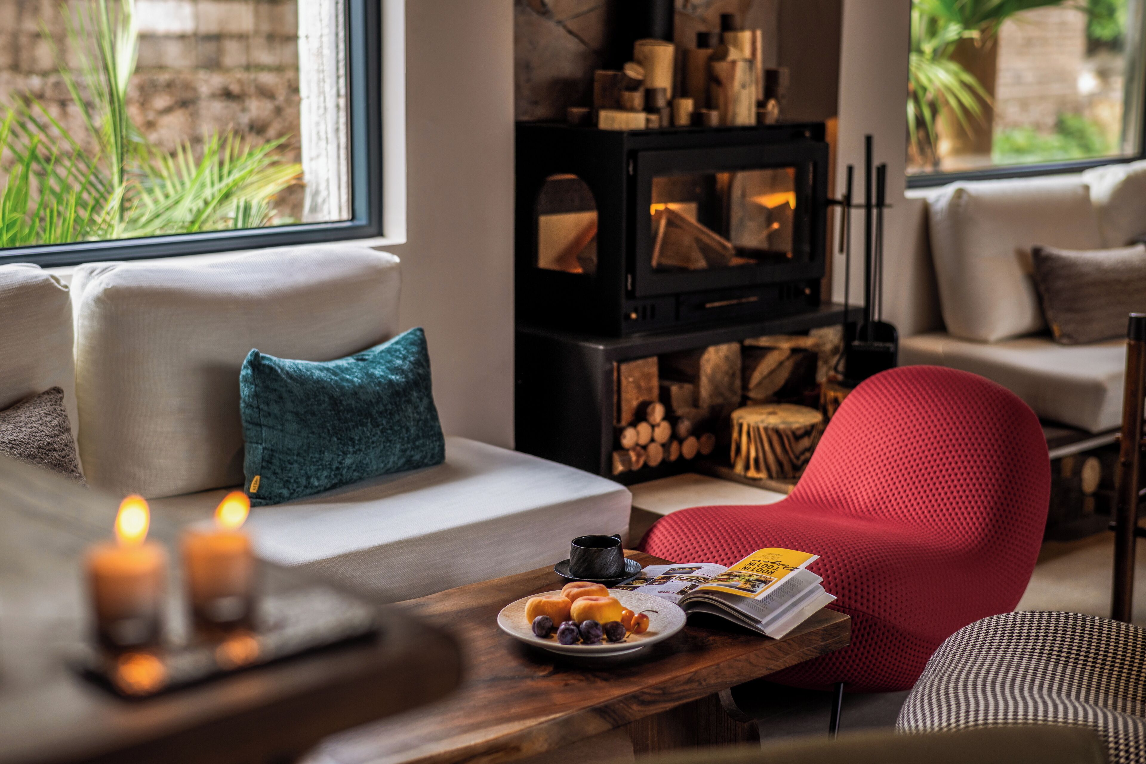 A cozy living room with a sofa, a red chair, a wooden table with a plate of food, a magazine, and a fireplace.