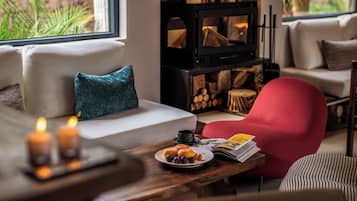 A cozy living room with a sofa, a red chair, a wooden table with a plate of food, a magazine, and a fireplace.