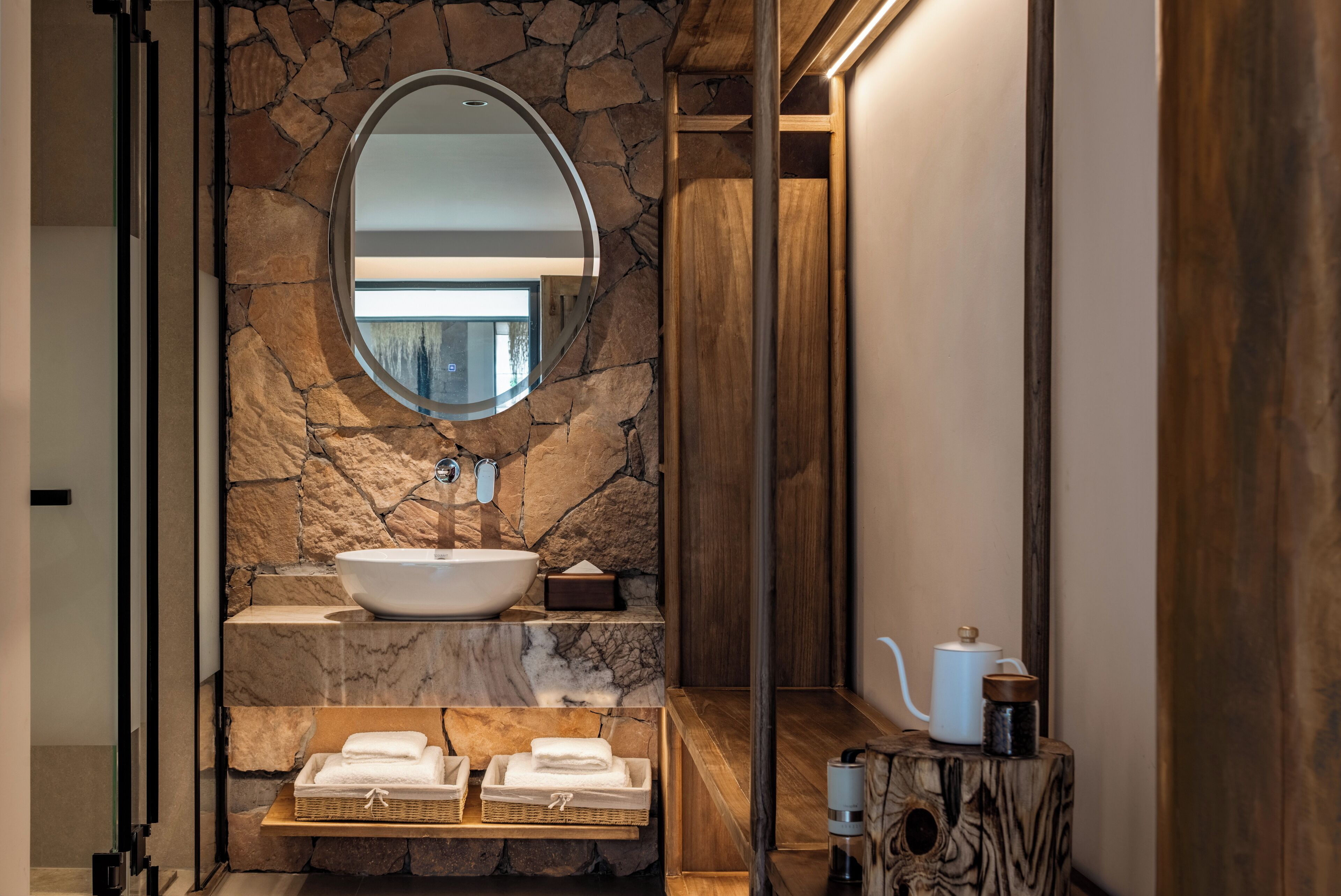 A bathroom with a stone wall, a round mirror, a white sink, a wooden shelf with towels, and a wooden cabinet with a teapot.