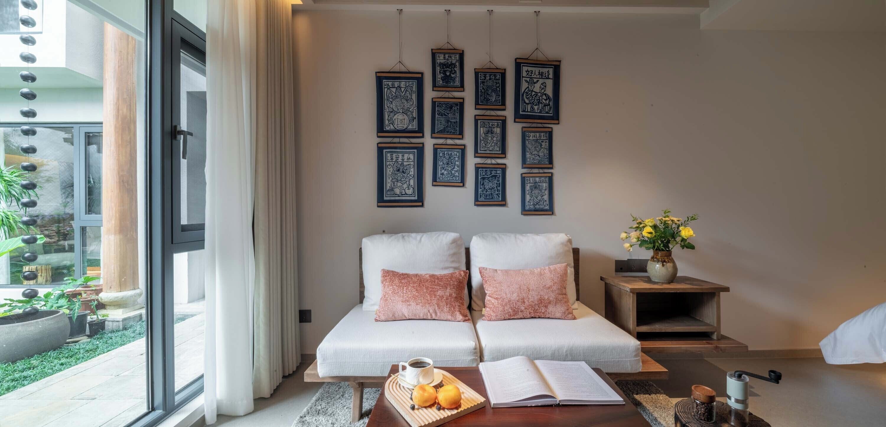 A modern hotel room with a large bed, a coffee table with breakfast items, a bookshelf, and a view of the garden through the window.
