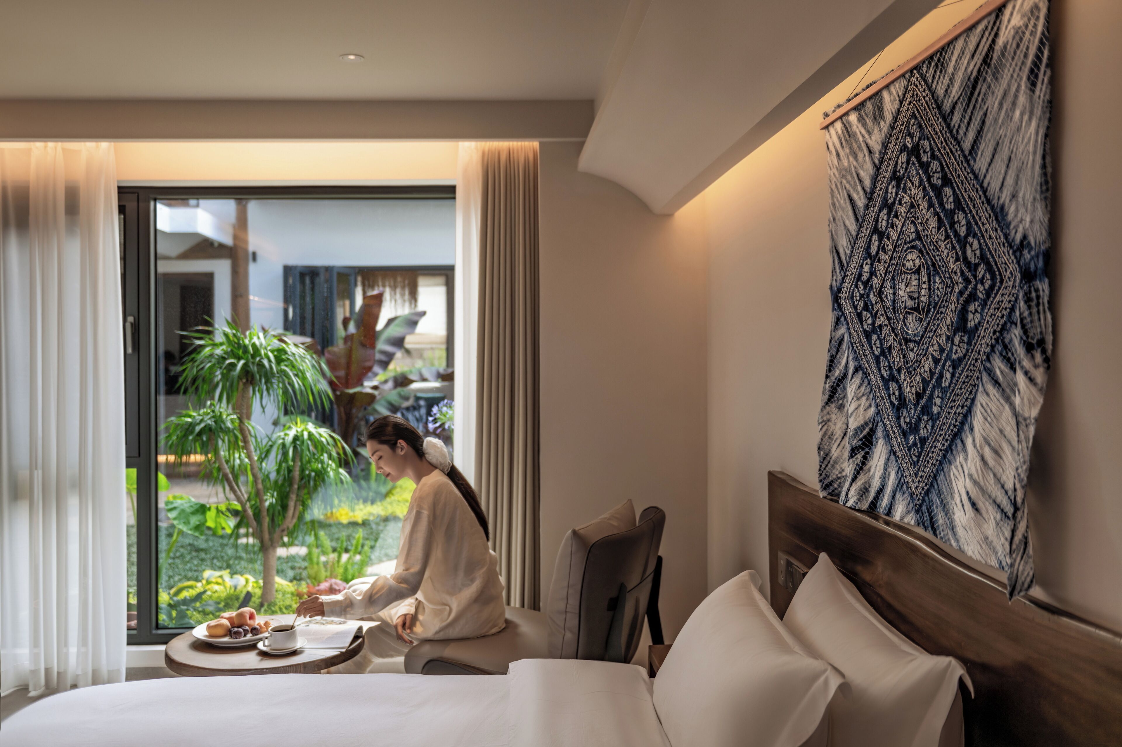 Quarto de hotel com cama, mesa de jantar com café da manhã, vista para um jardim e uma grande tapeçaria na parede.