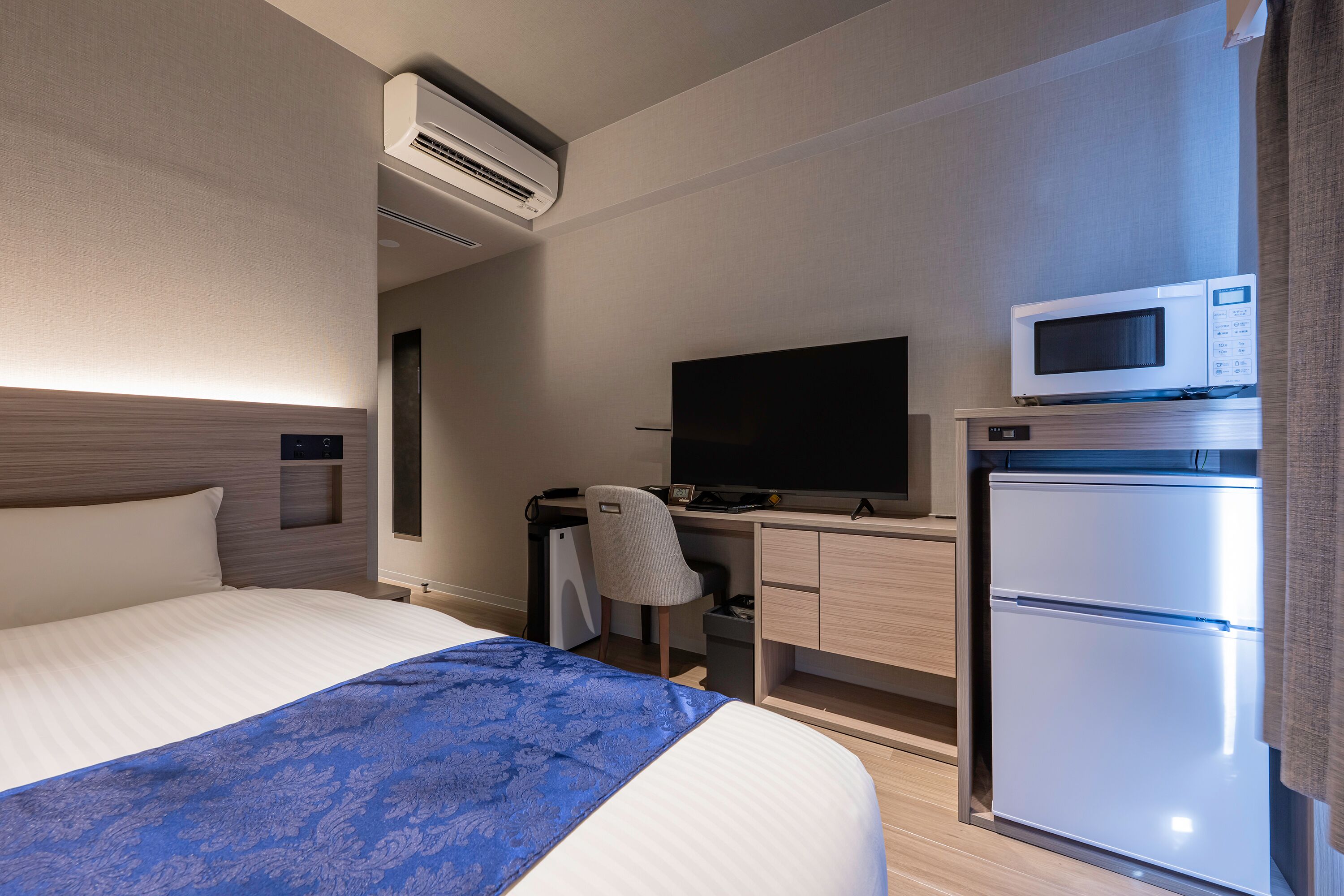 A hotel room with a bed, a desk with a television, a refrigerator, and an air conditioning unit.