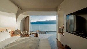A hotel room with a large bed, a TV, a seating area with a table, and a view of the sea and mountains.