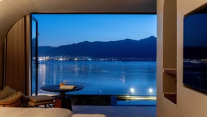 A room with a large window overlooking a body of water and mountains at dusk.