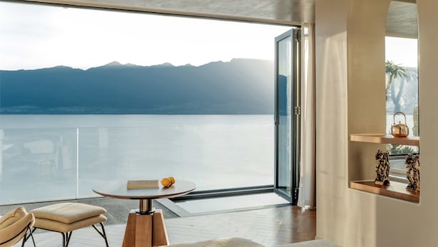 A modern room with a large glass door leading to a balcony overlooking a lake and mountains.