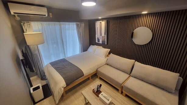 A modern hotel room with a bed, sofa, bedside table, lamp, and window with curtains. - GuestHouseAQUA203 (Hiroshima)