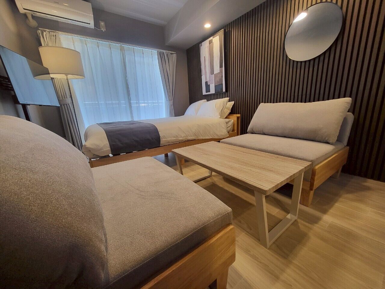 A modern hotel room with a bed, two sofas, a wooden table, and a large mirror.