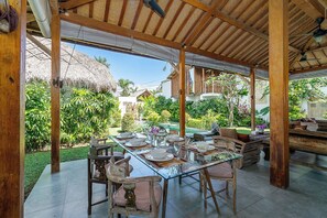 An outdoor dining area with a glass table and wooden chairs, surrounded by lush greenery and a thatched-roof structure.