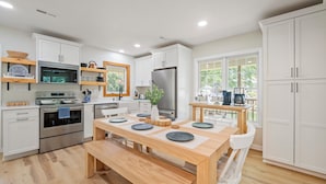 Una cocina moderna con armarios blancos, electrodomésticos de acero inoxidable y una mesa de comedor de madera dispuesta con platos y una banca.