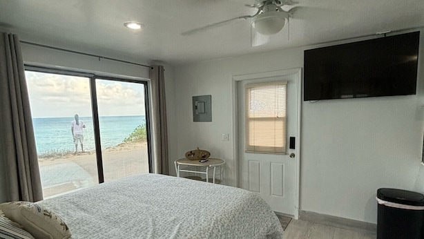A bedroom with a large bed, a ceiling fan, a TV mounted on the wall, and a view of the beach through the window. - Bimini Beach Getaway #2 (Alice Town)