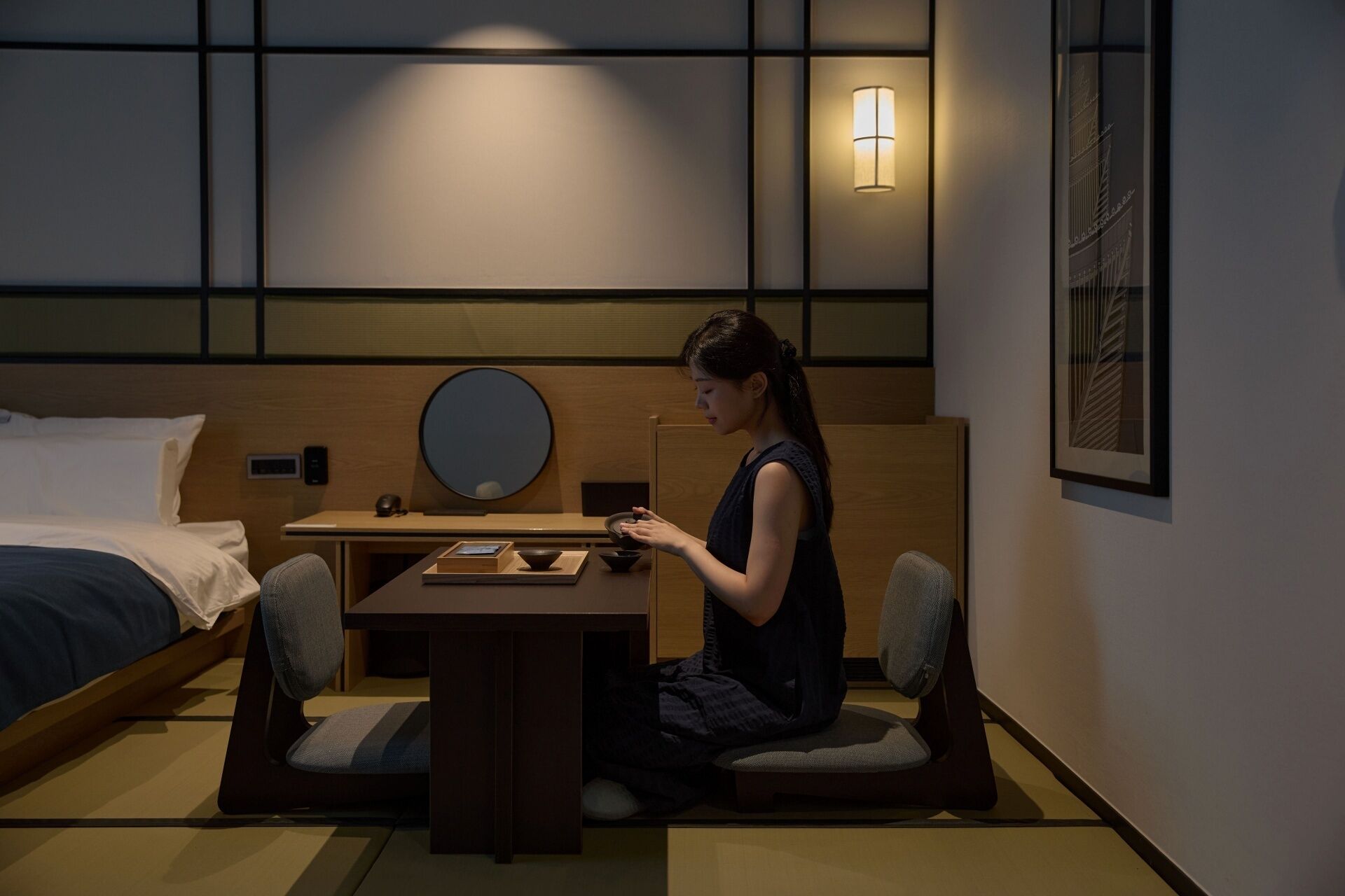 Une chambre d’hôtel comprenant un lit, un bureau avec une chaise, un miroir rond et un luminaire fixé au mur.