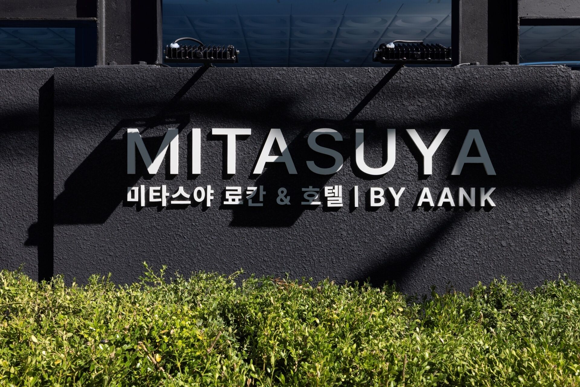 A sign for Mitasuya Iby Aank, a bank, with a dark wall and greenery in front.