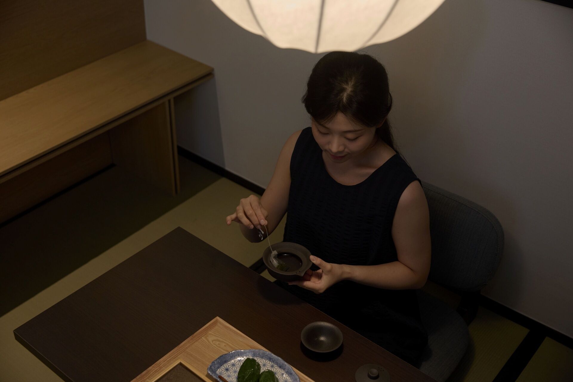 A person seated at a table with a teapot and a cup, a bowl, and a tray with a leaf.