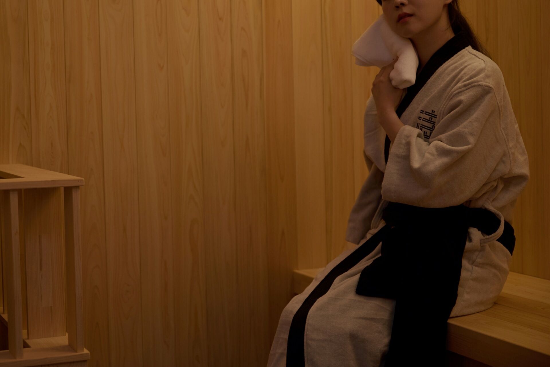 A person in a white robe with a black belt sitting on a wooden bench in a room with wooden paneling.