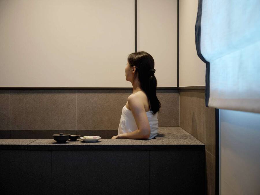 Una persona sentada en un banco en una habitación con pared de azulejos y una ventana con persiana blanca.