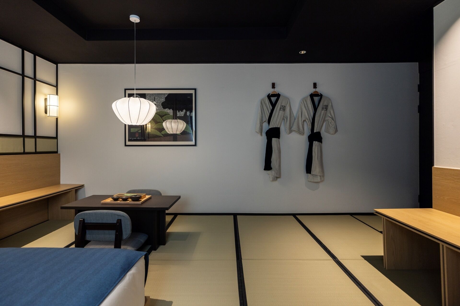 Ein modernes Zimmer mit Tatami-Böden, einem Holztisch, einer Pendelleuchte und zwei Kampfsport-Anzügen an der Wand.