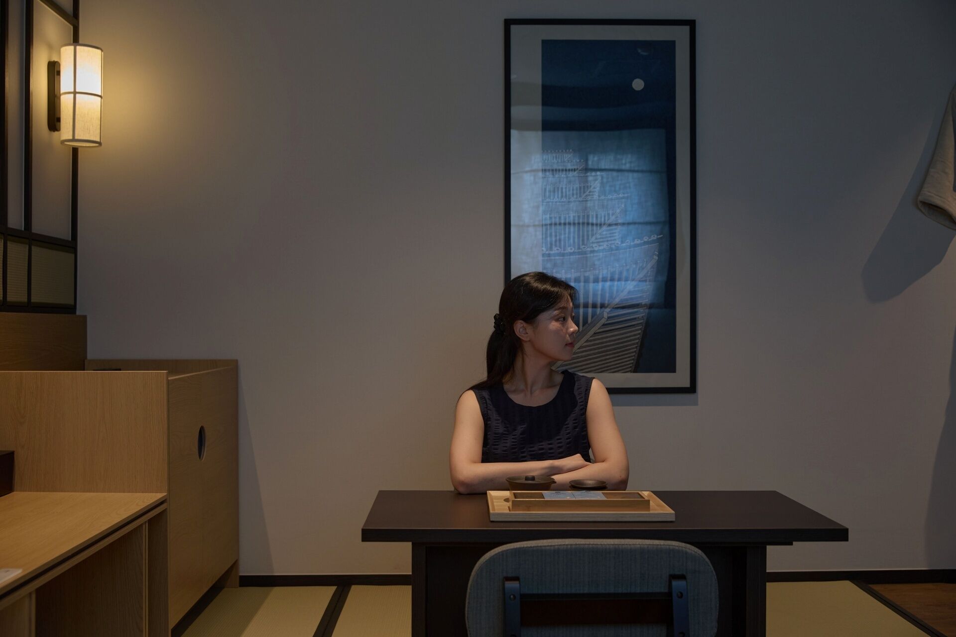 A person seated at a table in a room with a framed abstract painting on the wall and a wall-mounted light fixture.