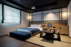 Una habitación de hotel moderna con cama, una mesita pequeña con silla, una mesita de noche y un ventanal con vistas.