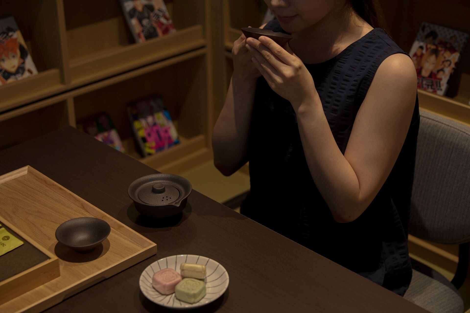 A person sitting at a table with a teapot, a cup, and a plate with sweets.