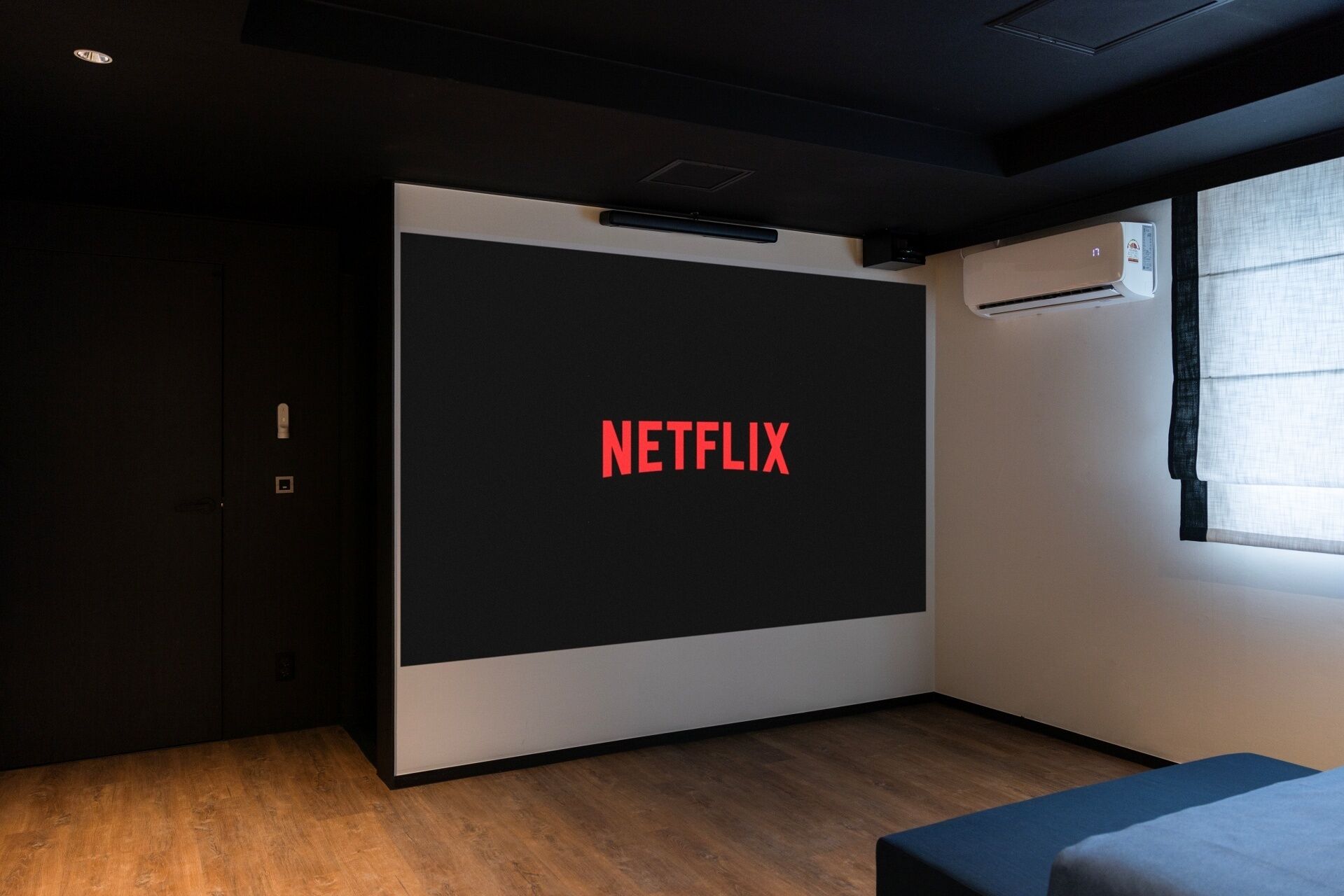 A modern room with a large screen displaying the Netflix logo, a dark door, and a window with blinds.