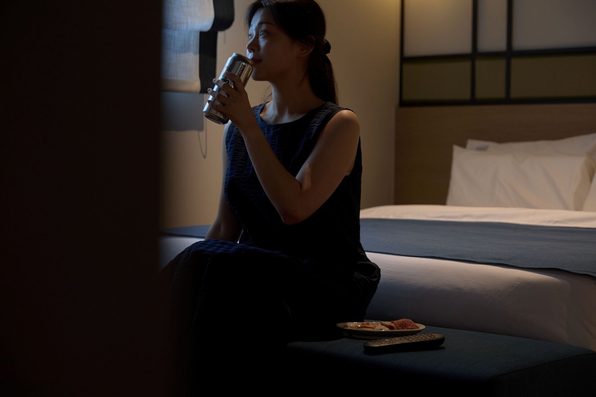 Una persona sentada en una cama en una habitación con poca luz, sosteniendo una lata. Un control remoto y un plato con comida están sobre una mesita de noche.