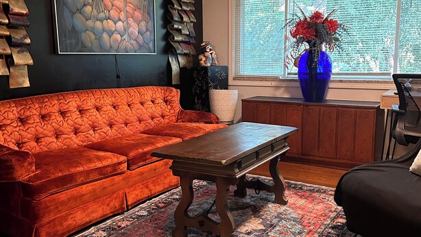 Una sala de estar con un sofá rojo, una mesa de centro de madera, un tapete estampado, un jarrón azul con flores y un cuadro grande en la pared.