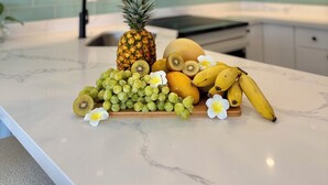 A kitchen counter with a fruit display including pineapple, kiwi, grapes, bananas, and flowers. - Wake Up to Waves Oceanview Retreat (Car Available) (Honolulu)