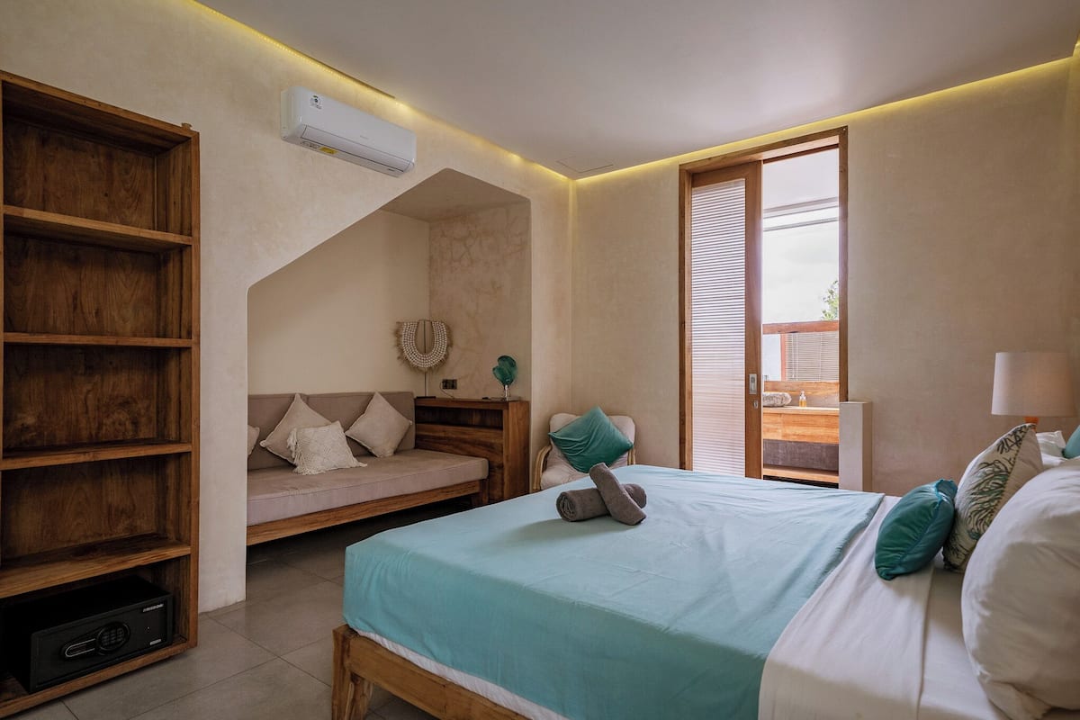 A bedroom with a bed, a wooden shelf unit, a sofa, and a window with blinds.