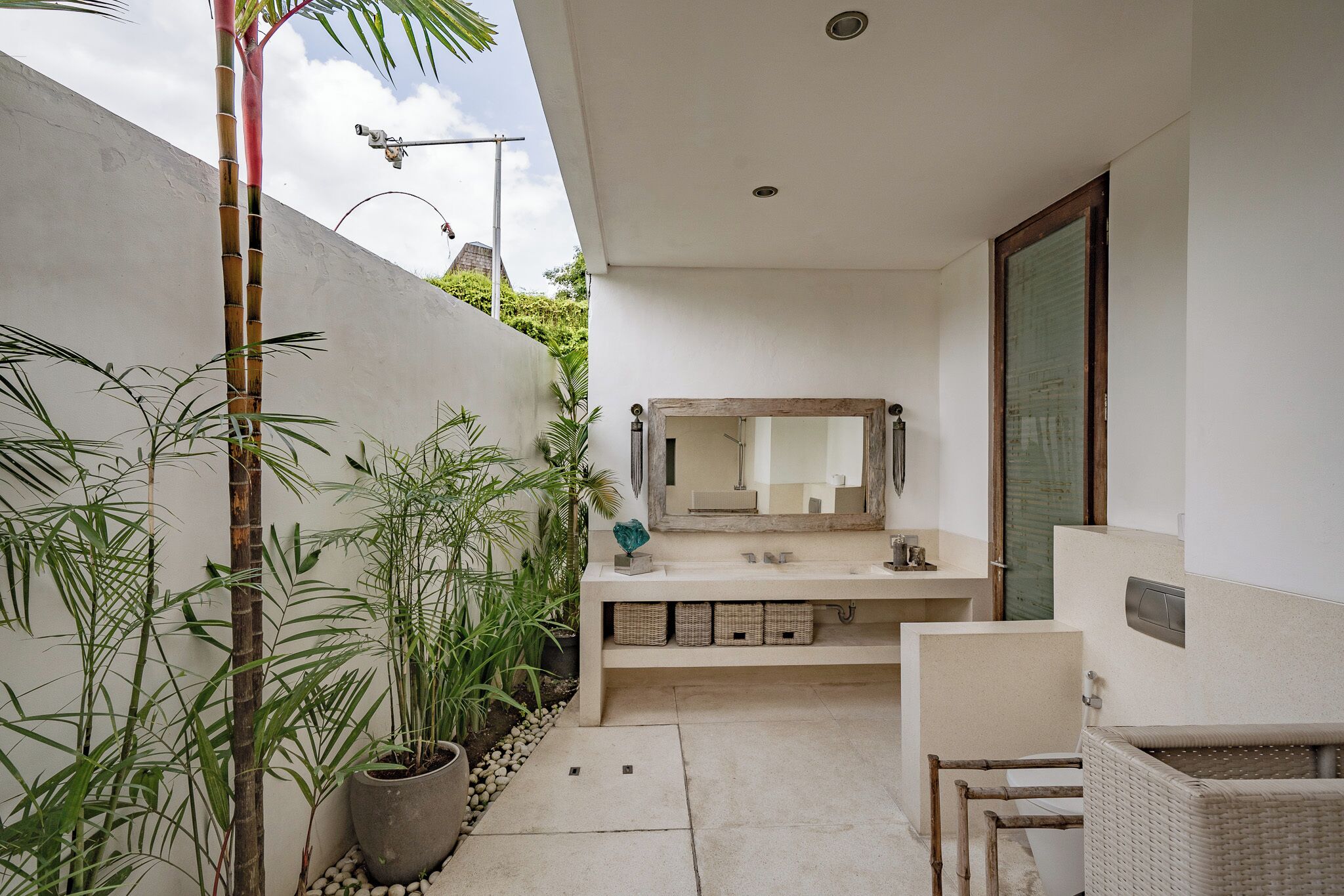 A modern bathroom with a large mirror, a sink, and a toilet. There are potted plants and a wicker chair.