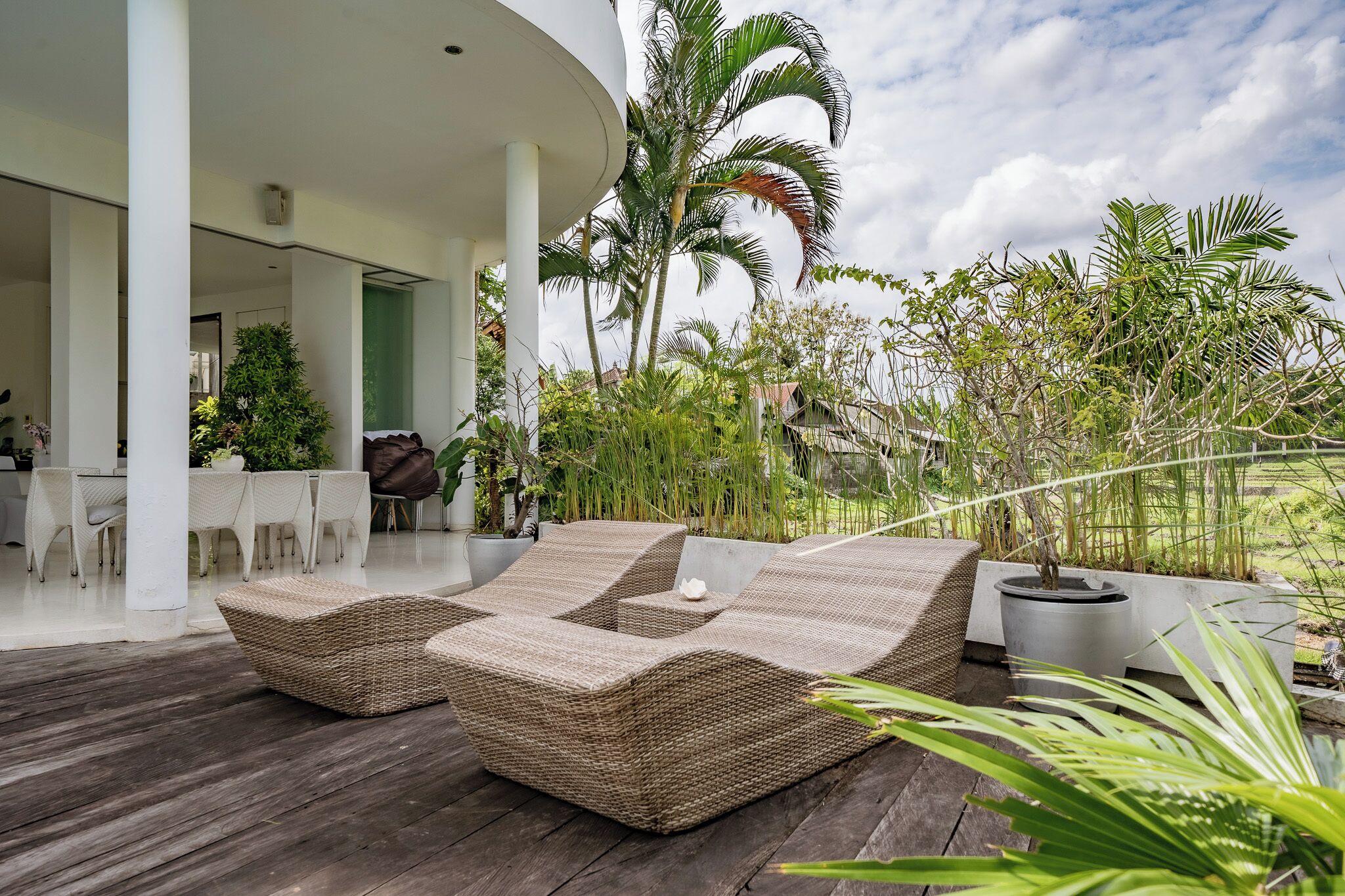 A covered outdoor seating area with wicker furniture, surrounded by lush greenery and palm trees.