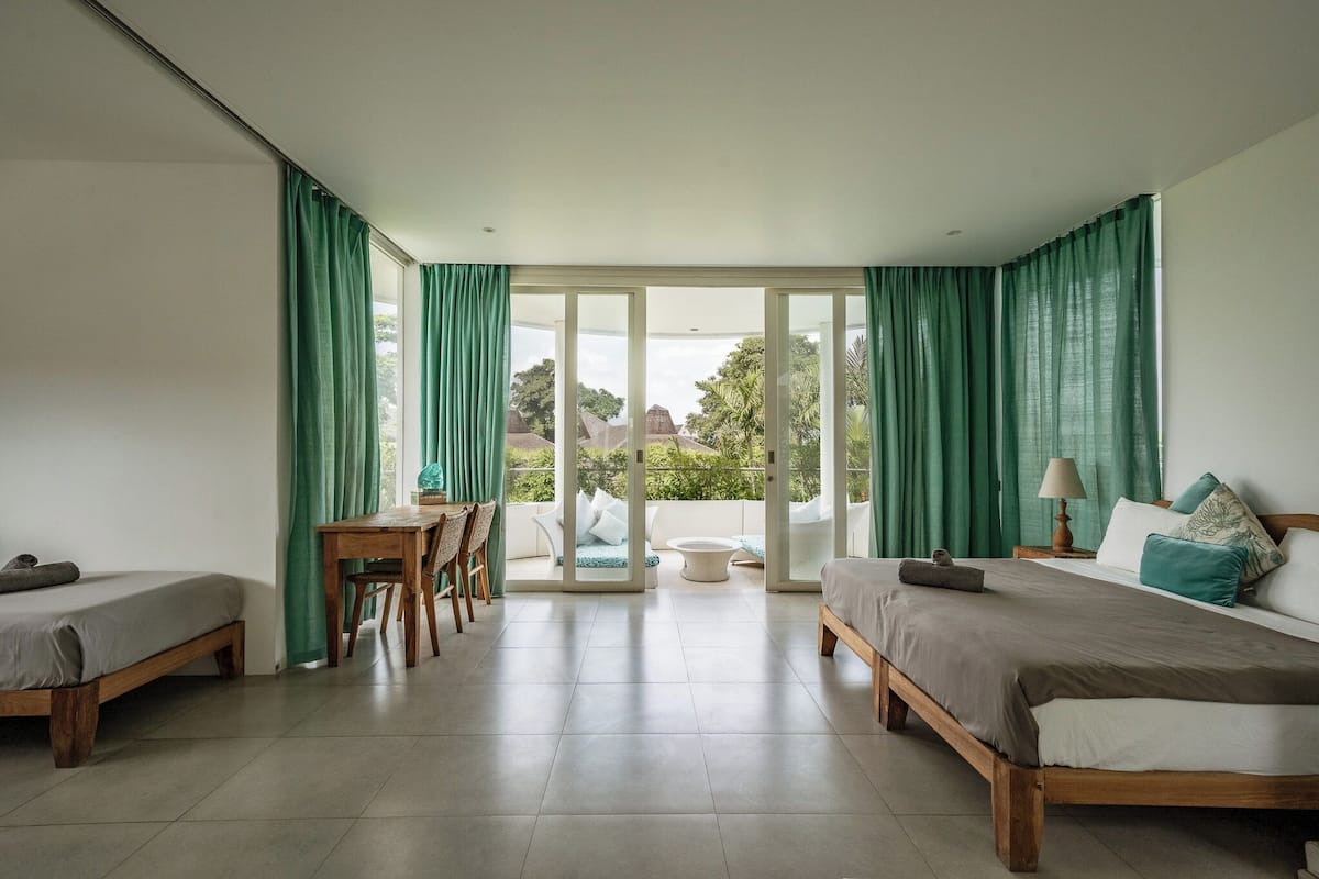 A room with two beds, a desk, and a balcony with green curtains.