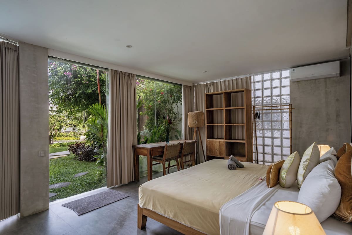 A bedroom with a large bed, a desk, and a chair. There is a view of a garden through the window.