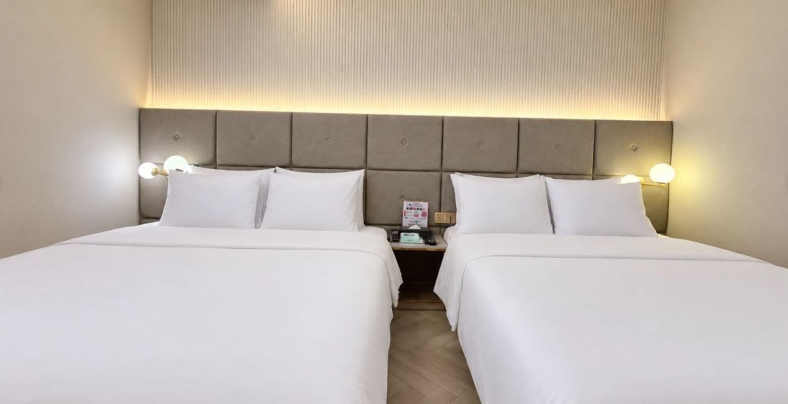 A hotel room with two beds, a headboard with integrated lighting, and a nightstand with a phone and a sign.