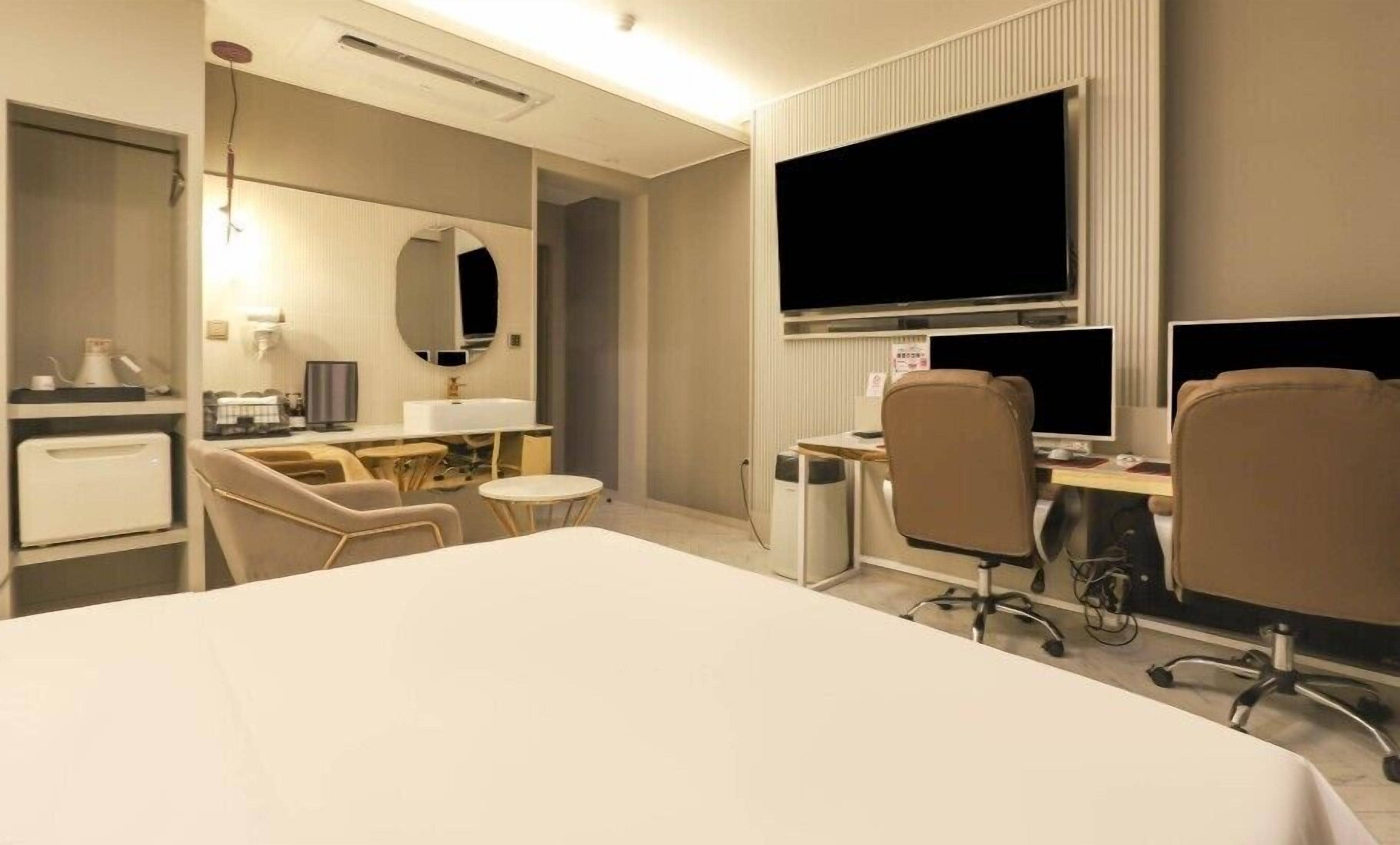 A hotel room with a bed, two chairs, a desk with a computer, a TV, and a small table with a lamp.