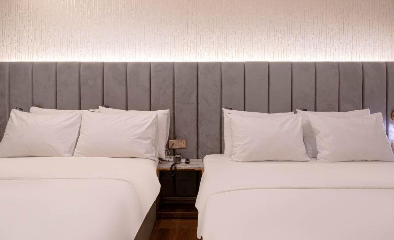 A hotel room with two beds, a headboard, and a bedside table with a lamp.