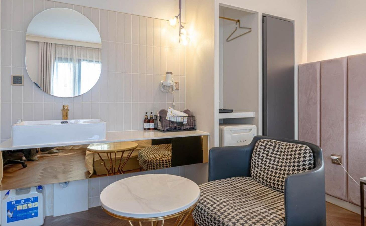A compact hotel room with a round mirror, a sink, a small table, a patterned armchair, and a wardrobe.