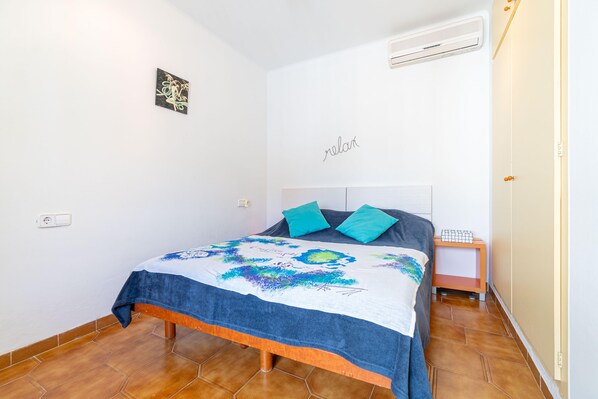 Un dormitorio con una cama, una mesita de noche, aire acondicionado de pared y un estante con objetos.