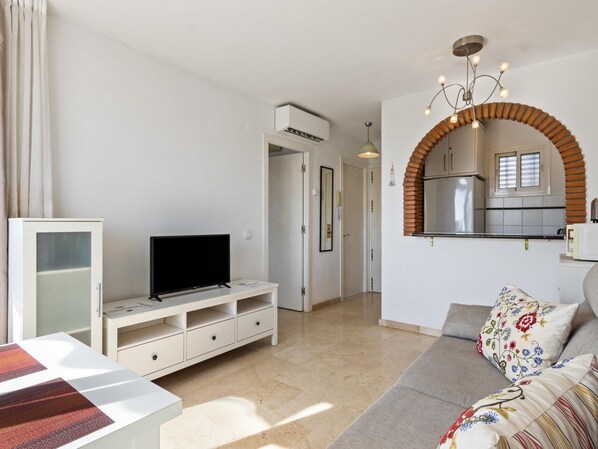 A modern living room with a flat-screen TV on a white TV stand, a sofa with floral cushions, a kitchen with a brick arch, and a hallway leading to a door.