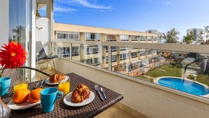 Ein Balkon mit Frühstücksangebot, darunter Croissants, Orangen und Saft, mit Blick auf einen Swimmingpool und ein Gebäudekomplex.
