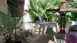 Un patio con mesa y sillas de madera, rodeado de macetas con plantas y un muro de piedra.