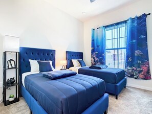 A room with two beds, a nightstand, a shelf with decorative items, and a window with an ocean-themed curtain.