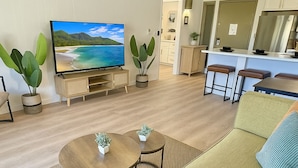 A modern living room with a large flat-screen TV, a sofa, two coffee tables, and a kitchen area with a refrigerator and bar stools. - Ocean view A/C, pool/hot tub coconut marketplace (Kapaʻa)